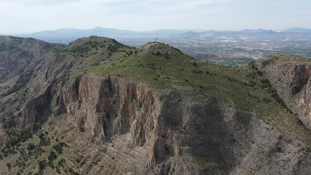 View of the wide mountainous landscape of Spain to the west. The drone is flying towards the summit cross of Mount Muela near the city of Orihuela and stops at the cross.