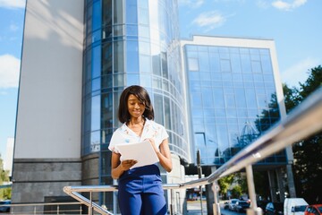 Fototapeta premium Portrait of young female African American job seeker keeping a folder with CV in her hands standing against office building. Blurred background with copy space