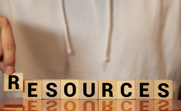 Businessman Protecting His Resources Holding His Hands Above A Row Of Wooden Blocks With The Word - Resources - Sheltering Them From Harm Or Over Exploitation.