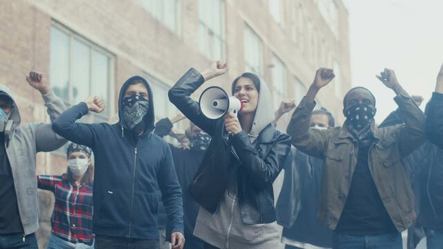 Caucasian young woman in hood screaming mottos in megaphone. Multiethnic male and female crowd of protesters shouting slogans and protesting. Girl leading at manifestation for human rights.