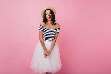 Debonair caucasian lady in long skirt posing with pleasure in studio. Pretty european woman in hat standing on pink background with charming smile.
