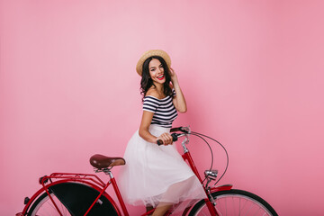 Excited latin girl sitting on bicycle with cheerful smile. Studio portrait of fashionable lady in lush skirt.