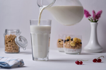 A man pours milk into a glass for breakfast. Muesli with yogurt and fresh berries served in a glass in the background. Breakfast on a white wooden table.