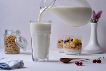 A man pours milk into a glass for breakfast. Muesli with yogurt and fresh berries served in a glass in the background. Breakfast on a white wooden table.