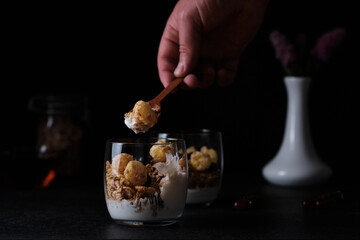 A man takes a spoon with granola and yogurt served in a glass for dinner. Black background around. Hands of a man in a frame.