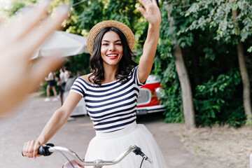 Obraz premium Excited girl sitting on bicycle and laughing to camera. Jocund latin female model waving hand on nature background.