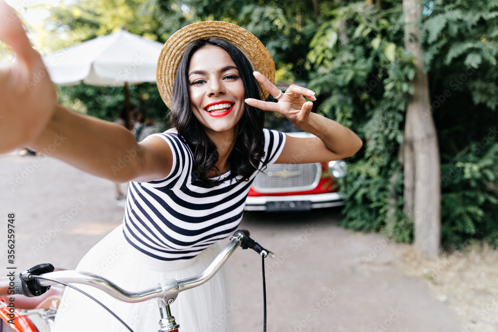 Wall mural positive white girl in straw hat making selfie in summer day. blithesome female model sitting on bic