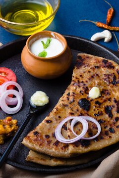 Aloo Paratha / Indian Potato Stuffed Wheat Bread served with Ghee (Indian Butter), Curd & Pickle, Tomato & Onion. Top View on a Dark Moody Background. Traditional North Indian or Punjabi Breakfast.