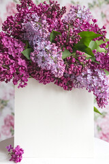 White sheet in beautiful blooming lilac on a white stand on a floral background. Greeting card, place for text, mock up