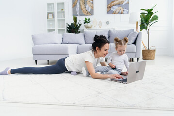 Young mother and her adorable daughter are using laptop while lying on the floor.