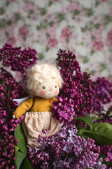 Little golden-haired angel in the blue, pink, purple, violet lilac flowers. Handmade toy in violet lilac colors. Greeting card.