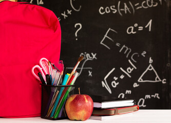 Coloured school supplies, apple, books, backpack on a white contertop on chalkboard background with formulas.