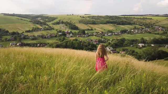 Yong woman with long hair in red dress walking in summer field with tall green grass on rural hill environment.