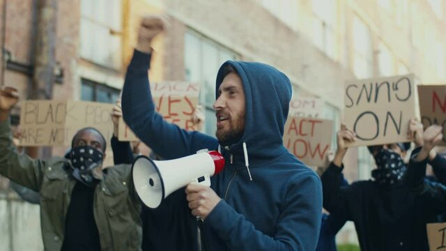 Caucasian young handsome guy protesting in middle of multiethnic crowd of protesters and screaming mottos in megaphone. Guy leading at manifestation for human rights.