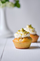 Fruit cupcakes with cream, decorated with ground pistachio and flowers in a vase on a white wooden table. Morning, rustic still life.