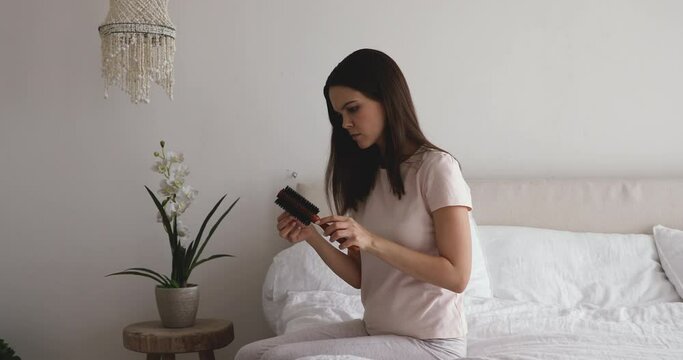 Serious young girl brushing hair with round hair tool, worrying about hair loss shedding or bad condition. Upset millennial woman sitting on bed, styling hair with comb, hormonal imbalance concept.