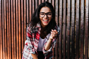 Dreamy tanned female model posing emotionally on wooden background. Outdoor portrait of enchanting girl wears glasses.