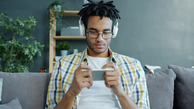 Slow motion of happy Afro-American guy playing video game in smartphone and enjoying music through headphones in modern apartment. People and devices concept.