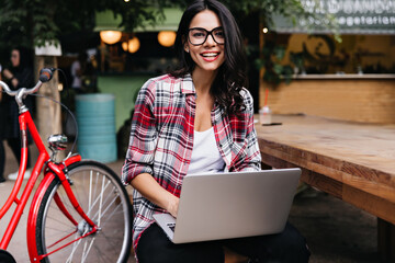 Laughing latin black-haired girl posing with bicycle and laptop. Happy stylish lady with computer sitting on the street.