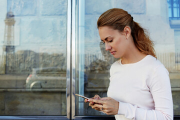 Fototapeta premium Young charming Caucasian female with trendy look use mobile phone while waiting taxi on bus stop,