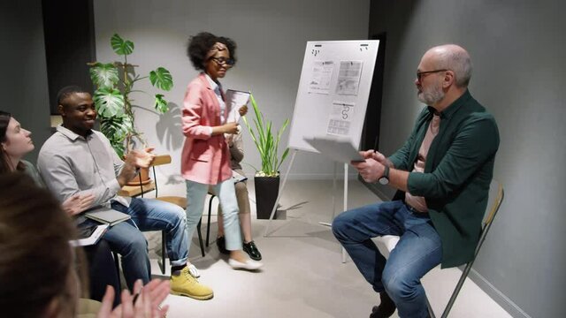 Senior male business coach introducing young African American female colleague to audience. Cheerful woman standing up, smiling and giving a speech to team of office workers