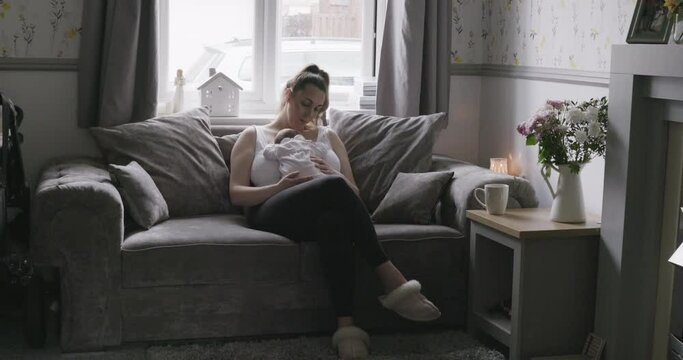 New mother sitting on sofa in living room, holding and looking at her newborn baby daughter.