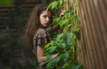 A beautiful, slender girl in a summer sundress poses against a wooden lattice fence with ivy