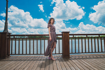 A slender, beautiful girl in a summer sundress against the background of a beautiful cloudy sky and a green river bank