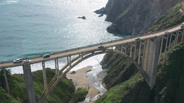 Aerial view of the picturesque arch bridge standing over the canyon among the beautiful hills covered with green grass. Cars move across the bridge. Famous landmark of USA under the cloudy sky. 4K