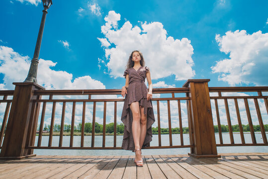 A slender, beautiful girl in a summer sundress against the background of a beautiful cloudy sky and a green river bank