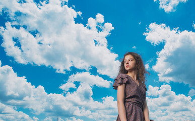 Fototapeta na wymiar A slender, beautiful girl in a summer sundress against the background of a beautiful cloudy sky and a green river bank