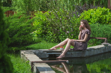 A slender, beautiful girl in a summer sundress sits on the green shore of the lake and eats strawberries
