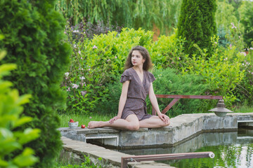 A slender, beautiful girl in a summer sundress sits on the green shore of the lake in a bowl of strawberries