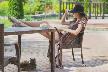 A slender, beautiful girl in a summer sundress and hat is sitting at a table in a summer cafe with her feet on the table