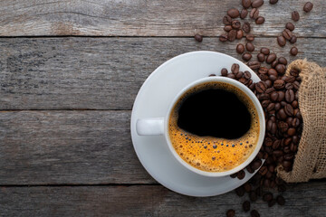 Top view above of Hot fresh black coffee with milk foam in a white ceramic cup with coffee beans roasted in sack bag on wood table background. Flat lay with copy space.
