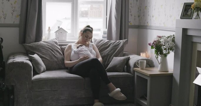 New mother sitting on sofa in living room, holding and looking at her newborn baby daughter.