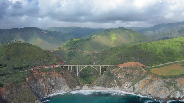 Amazing aerial nature view of the high green mountains along the deep-blue ocean coastline on a cloudy day. 4K drone footage of the arch bridge on Pacific highway running through the Big Sur Park, USA