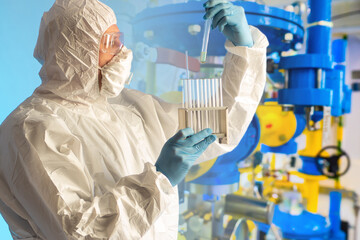 Work as a chemist in a dangerous production. Laboratory assistant in a protective suit with test tubes on the background of chemical laboratory equipment. Laboratory control of the production process.