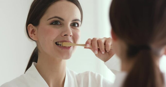Mirror reflection head shot close up young beautiful woman brushing teeth with bamboo wooden ecofriendly toothbrush, taking care of environmental pollution, enjoying morning healthcare routine.