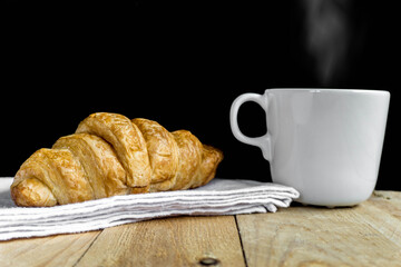 Fresh croissant on white fabric and hot coffee. Wooden table and black background with copy space