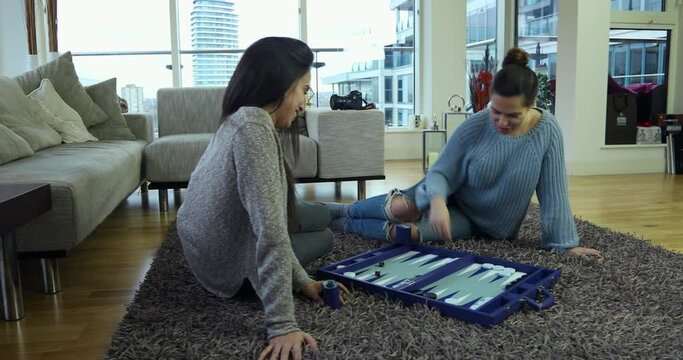 Two women playing backgammon seated on the floor of their city apartment. 