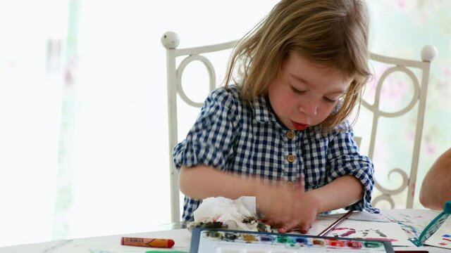 Footage of little girl with blond hair sitting indoors at a table, making a finger painting.