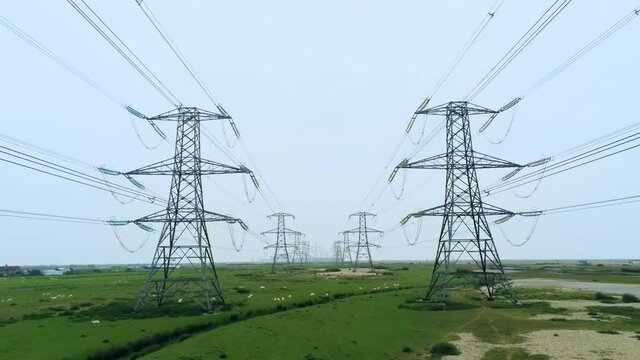 Moving through an avenue of power lines and pylons over green fields with sheep and a river on the horizon.