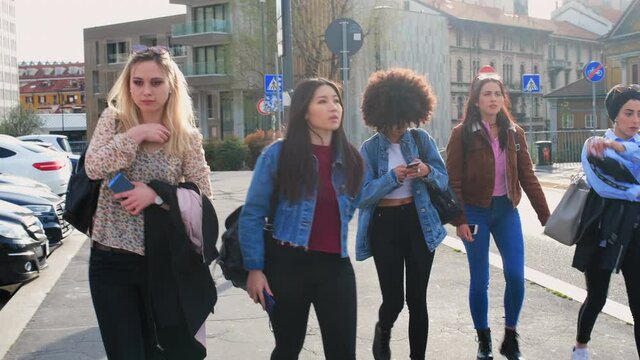 A group of girls, young women, female friends walking through a city, one on her phone.