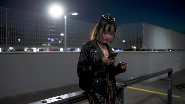 Young woman using her phone, looking at the screen adn using her thumb to text, in the city at night. 