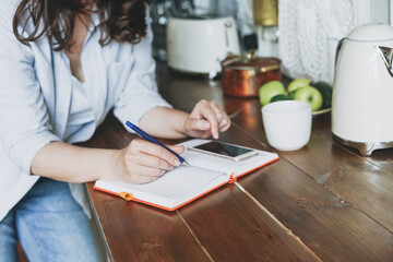 Fototapeta na wymiar Brunette woman in casual doing notes in daily book with and using mobile on kitchen at the home