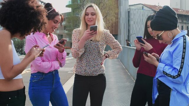A group of girls, young women, female friends standing using their phones, chatting, texting and laughing. 