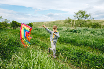 Cute white kid playing colorful kite outdoor at summer meadow or park. Color photography of caucasian young boy isolated at sunny blue sky and green field background