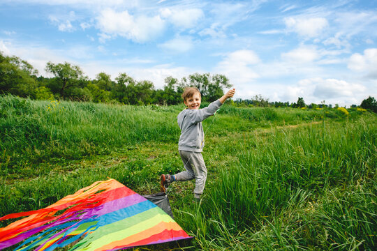 Cute white kid playing colorful kite outdoor at summer meadow or park. Color photography of caucasian young boy isolated at sunny blue sky and green field background