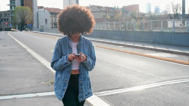 Girl with an afro in a denim jacket walking, concentrating on her phone. 